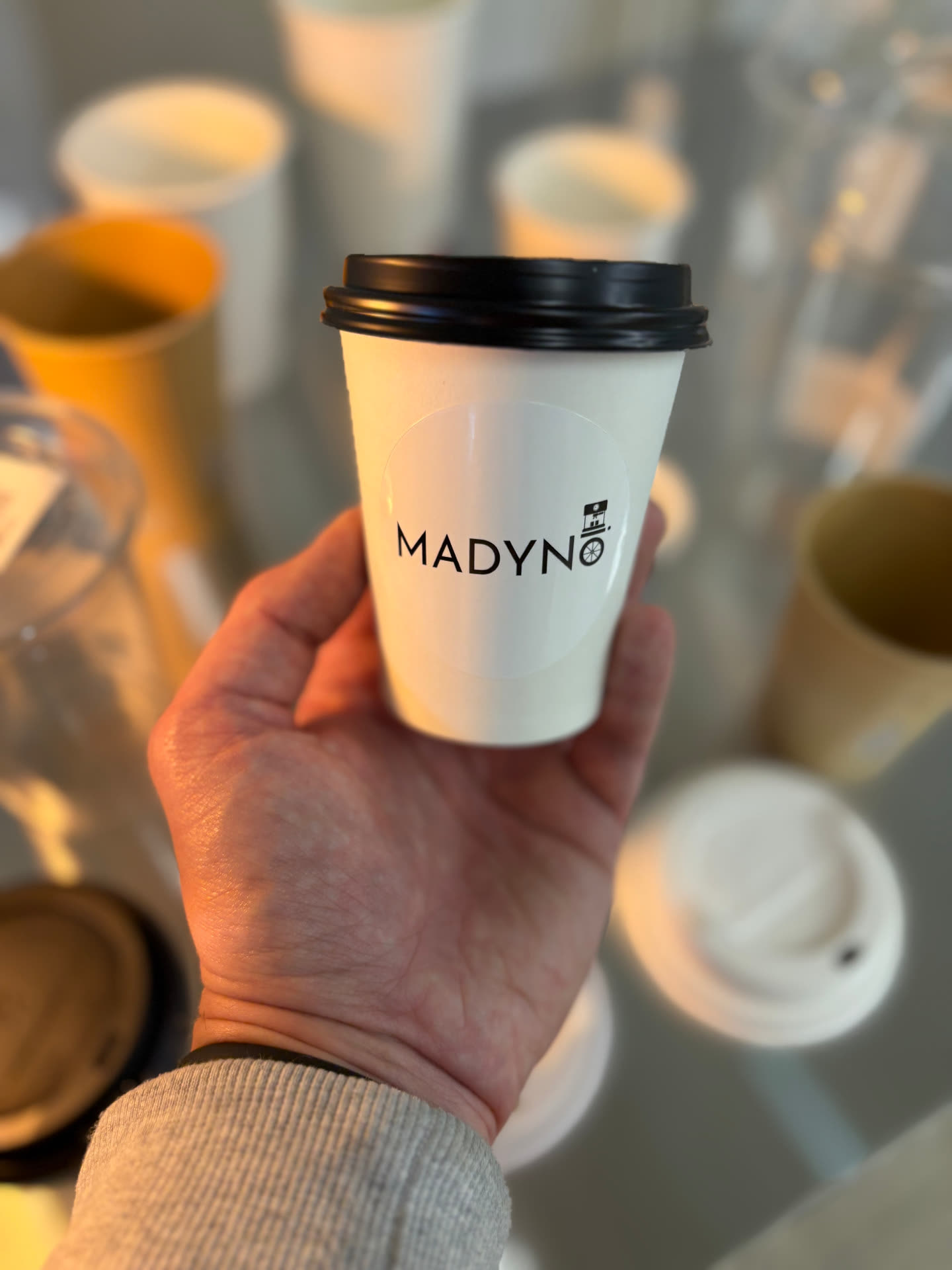 Weißer To-Go-Becher mit Madynio Logo-Etikett und schwarzem Deckel
